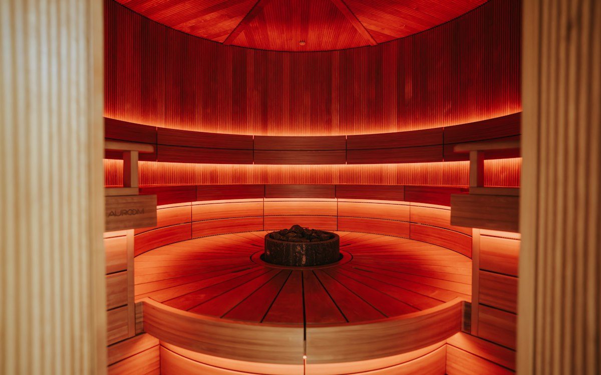A circular wooden sauna interior with red ambient lighting and a central stone heater