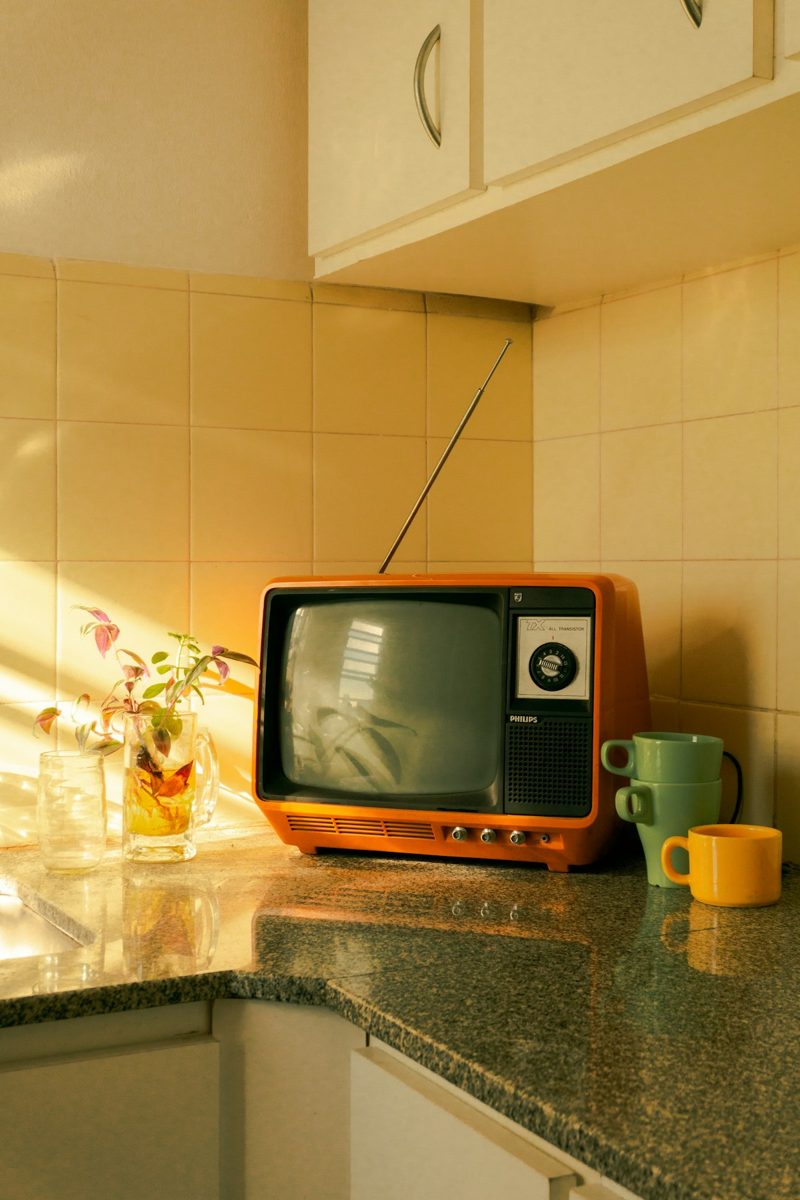 An orange vintage television on a granite kitchen counter with mugs and a plant